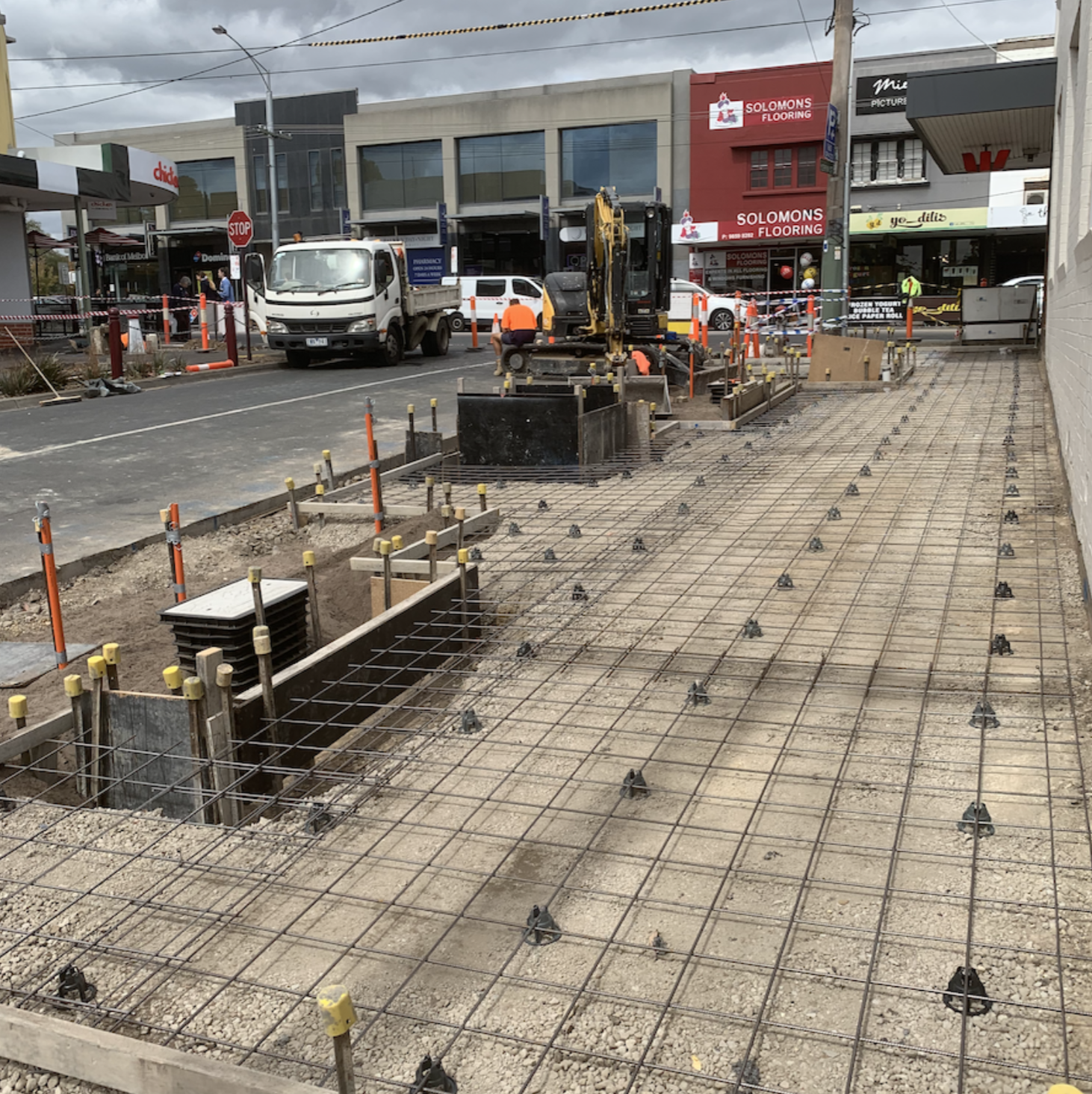 Construction site on a sidewalk with steel rebar grid, construction workers, and machinery, with stores and vehicles in the background.