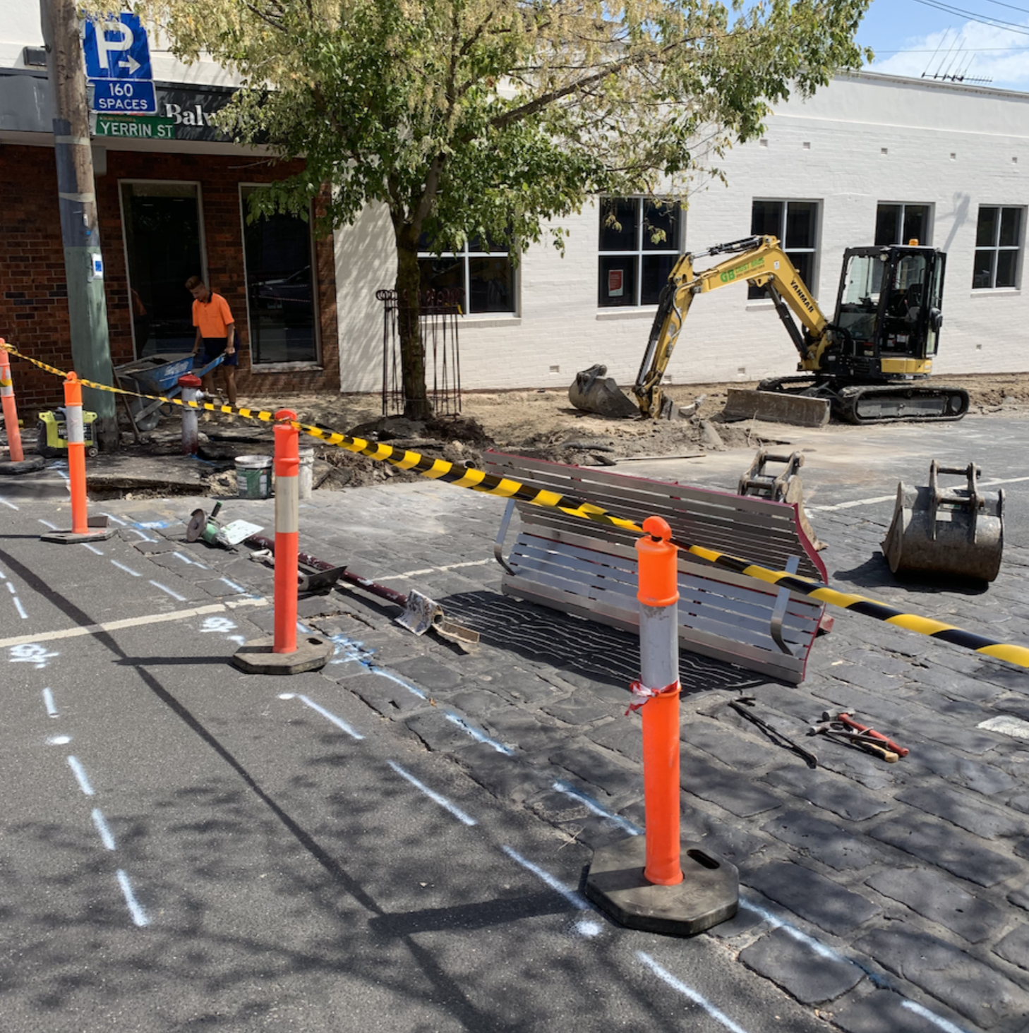 Street construction site with orange safety cones, yellow caution tape, a small excavator, and construction tools, with marked pavement and a building with white brick wall and trees in the background.
