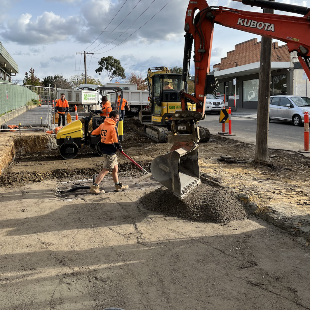 Construction workers and machinery working on a road repair site with a small excavator, a roller, and several workers in orange safety vests, in an urban area with parked cars and buildings visible in the background.