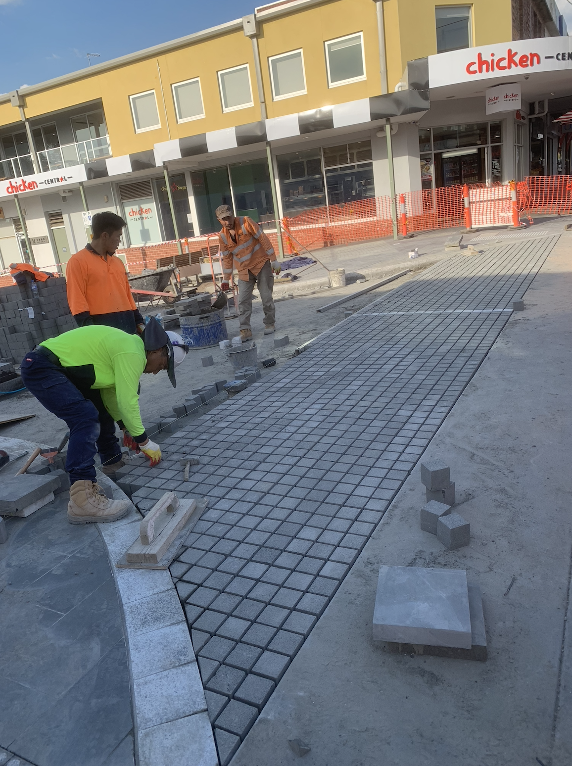 Construction workers laying paving stones on a sidewalk in front of a building with a yellow facade and signs that read 'chicken'.