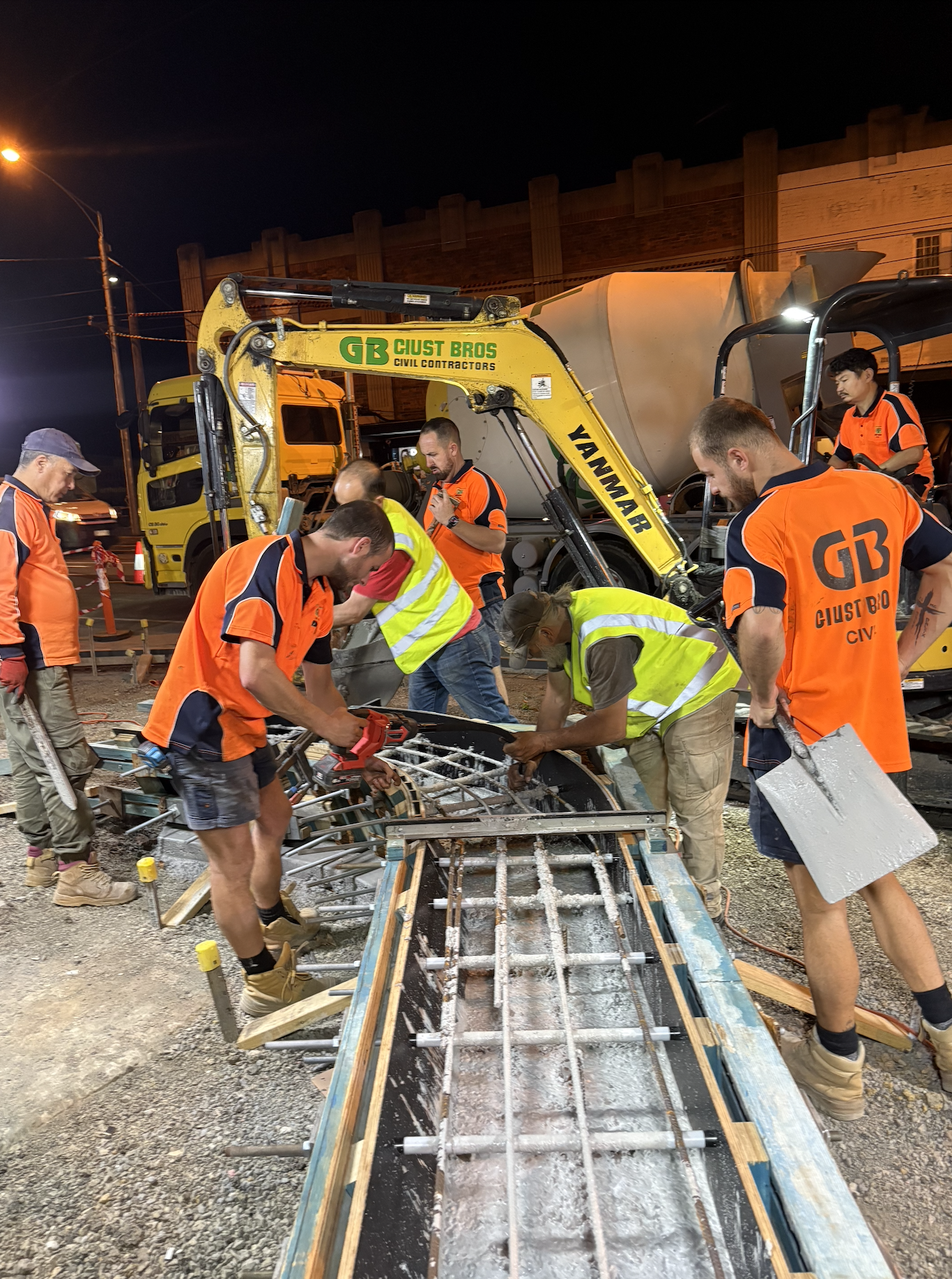 Construction workers in orange and yellow safety vests working together with machinery to reinforce a concrete structure at night.