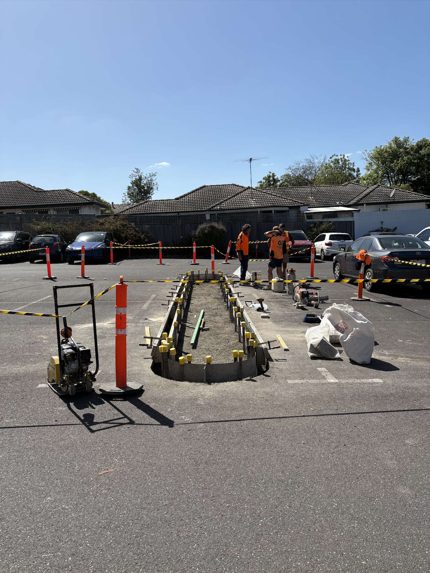 Construction workers installing a landscaped island in a parking lot, surrounded by orange cones and caution tape, with tools and materials scattered around.