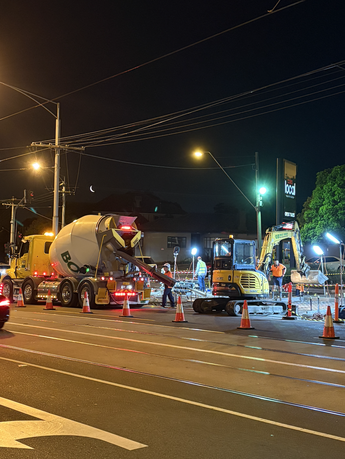 Construction workers operating heavy machinery at night on a city street, with a cement mixer truck, an excavator, and orange traffic cones, illuminated by bright work lights.