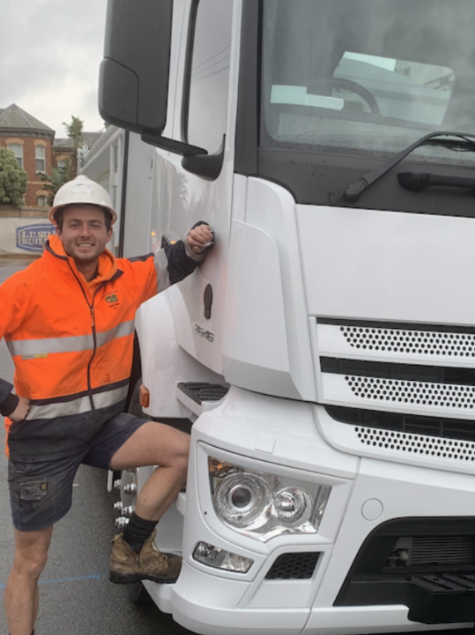 A man in an orange safety jacket and white hard hat leaning on a large white truck, smiling at the camera. He has one foot on the truck's step and is standing outdoors in a residential neighborhood.
