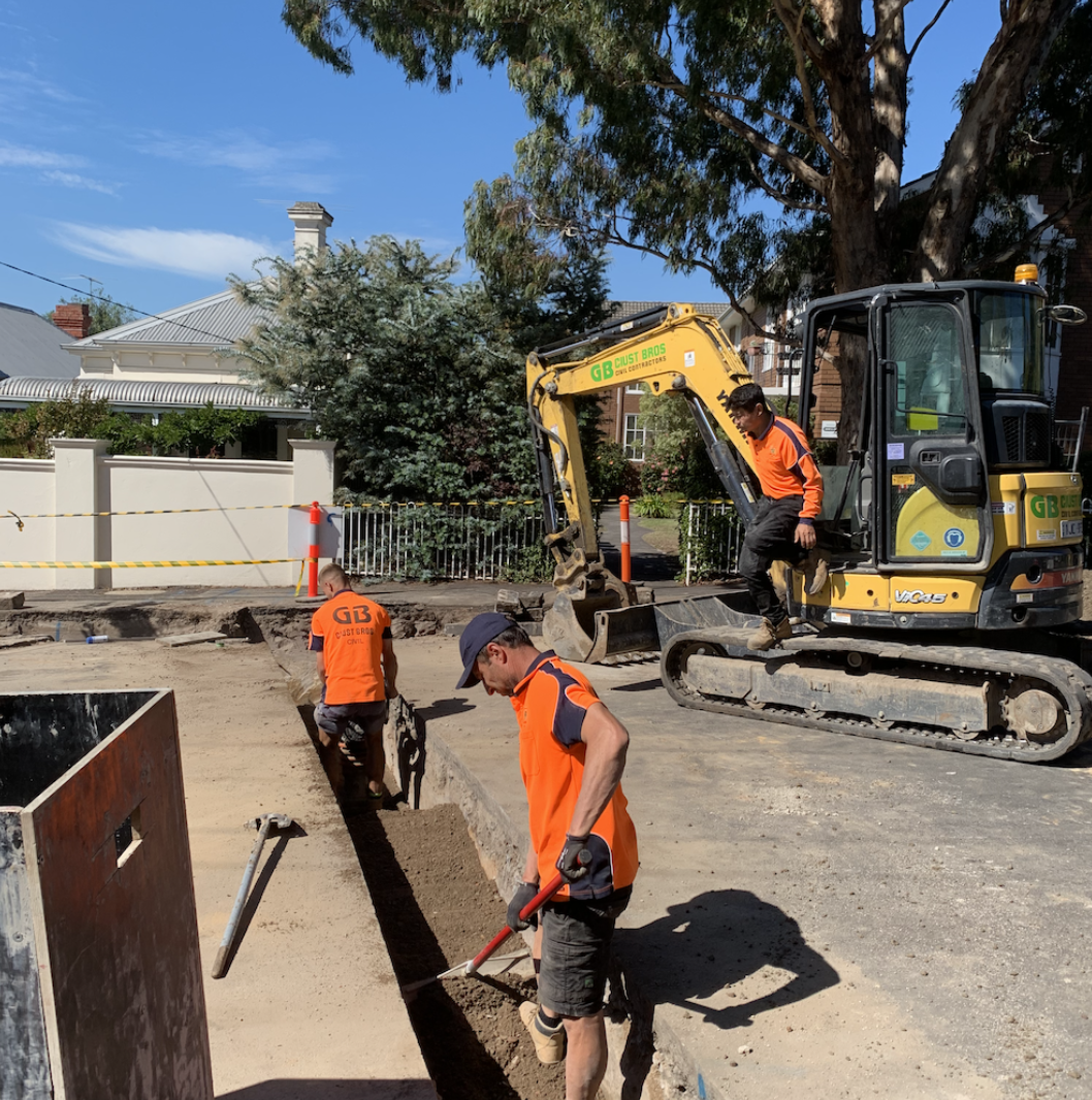 Construction workers in orange shirts working on a street excavation site, with small excavator and tools, surrounded by safety barriers and residential houses, during daytime.