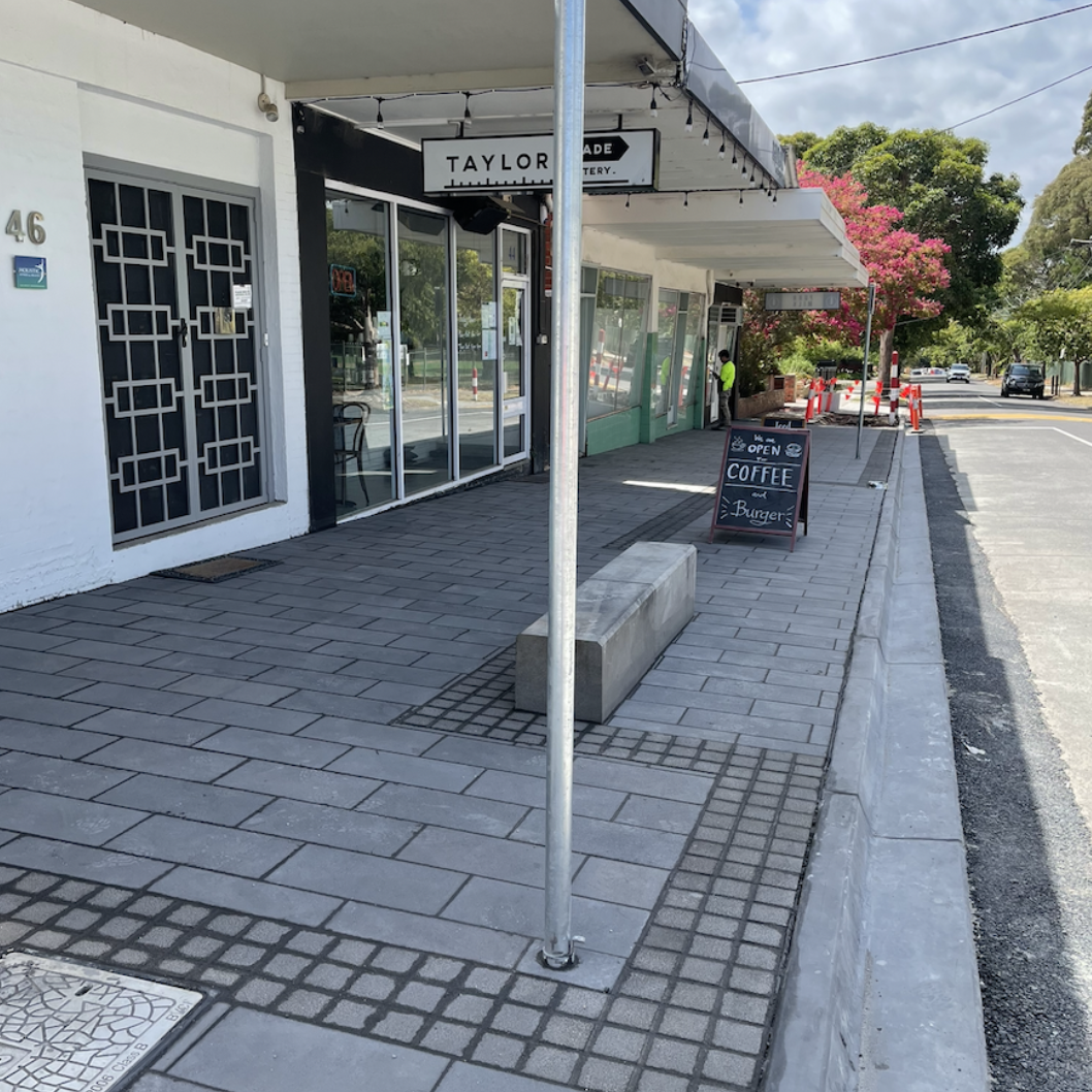 Sidewalk in front of a modern coffee shop with glass doors, a sidewalk sign advertising coffee and burgers, and a pedestrian on the sidewalk. There are trees with pink flowers and cars on the street.