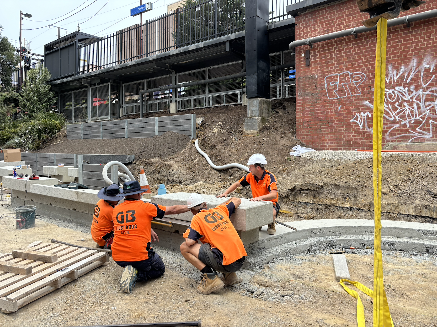 Four construction workers wearing orange shirts and white helmets are working on a concrete barrier at a construction site. They are handling and positioning the barrier, with tools and construction materials nearby. The background includes a brick wall with graffiti and an elevated sign indicating street name.