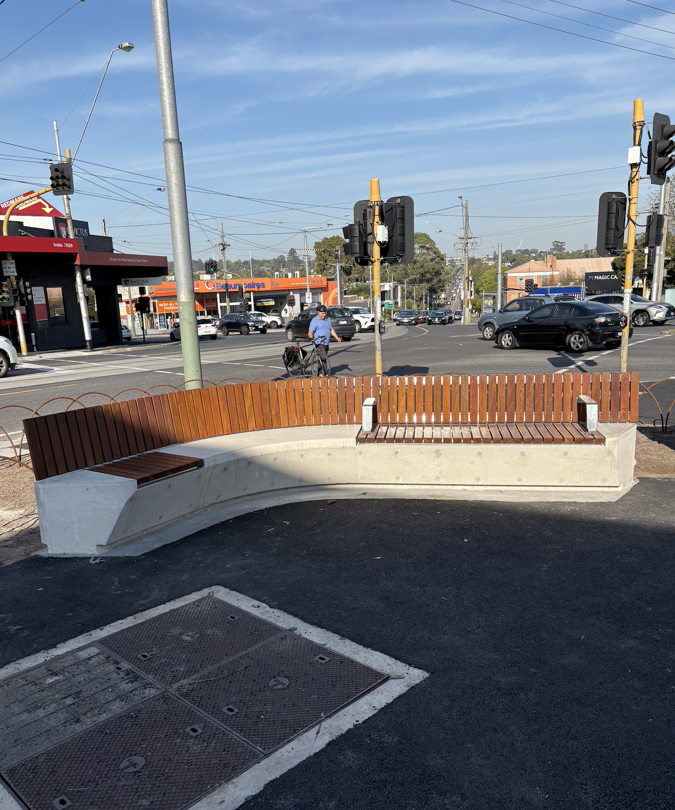Curved outdoor bench made of concrete with wooden slats in an urban setting, near a crosswalk and traffic lights, with stores and cars in the background.