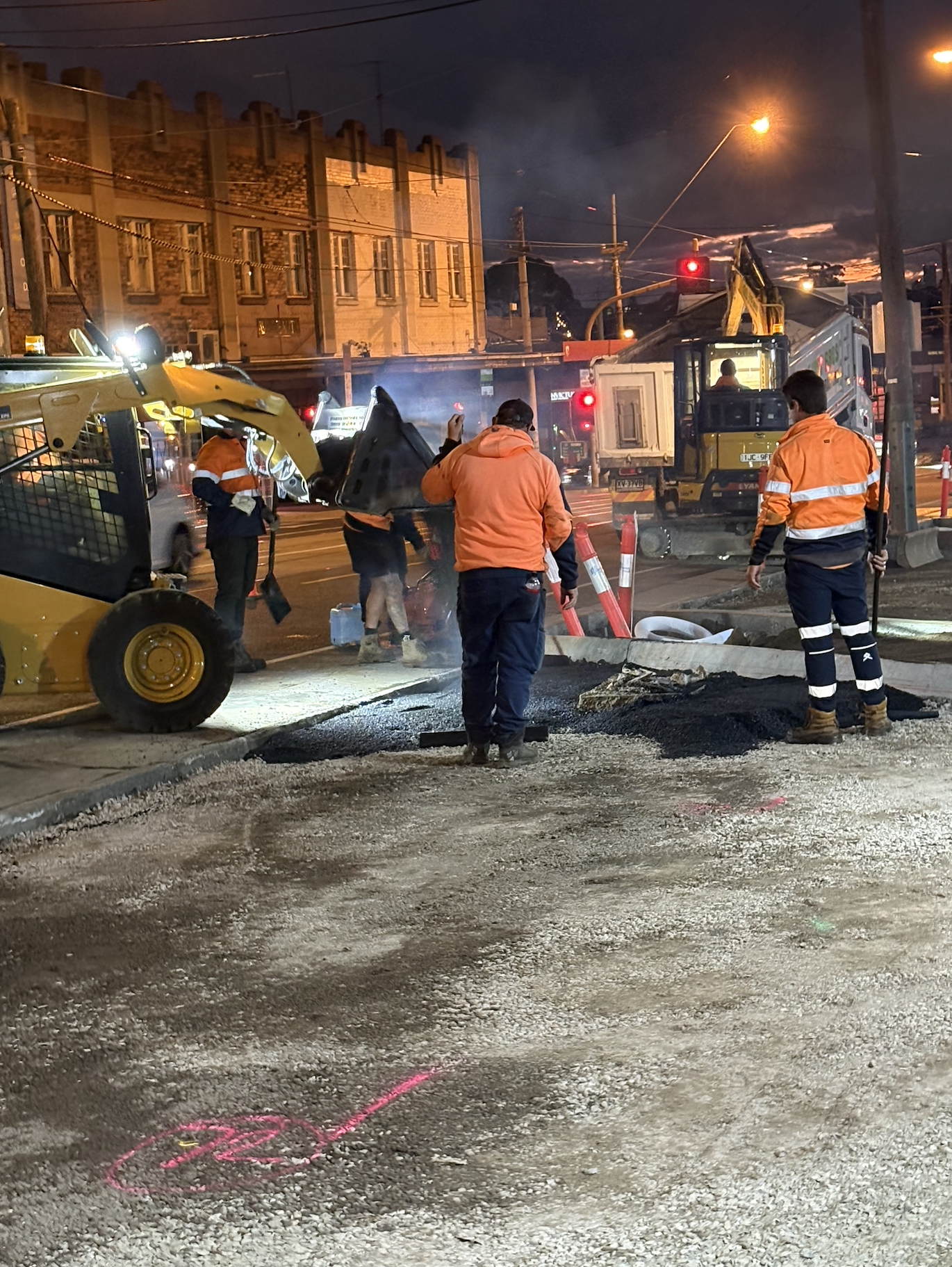 Construction workers in orange safety jackets working on a street at night, with construction equipment and traffic signals visible.
