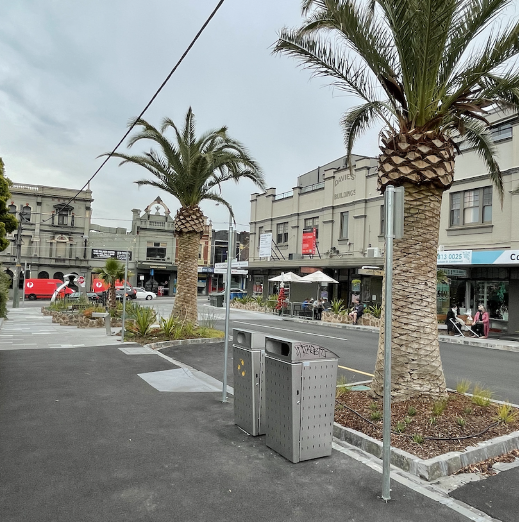 A street scene with two palm trees, trash cans, and a row of shops and cafes with outdoor seating. People are sitting and walking, and a red bus is visible in the background.