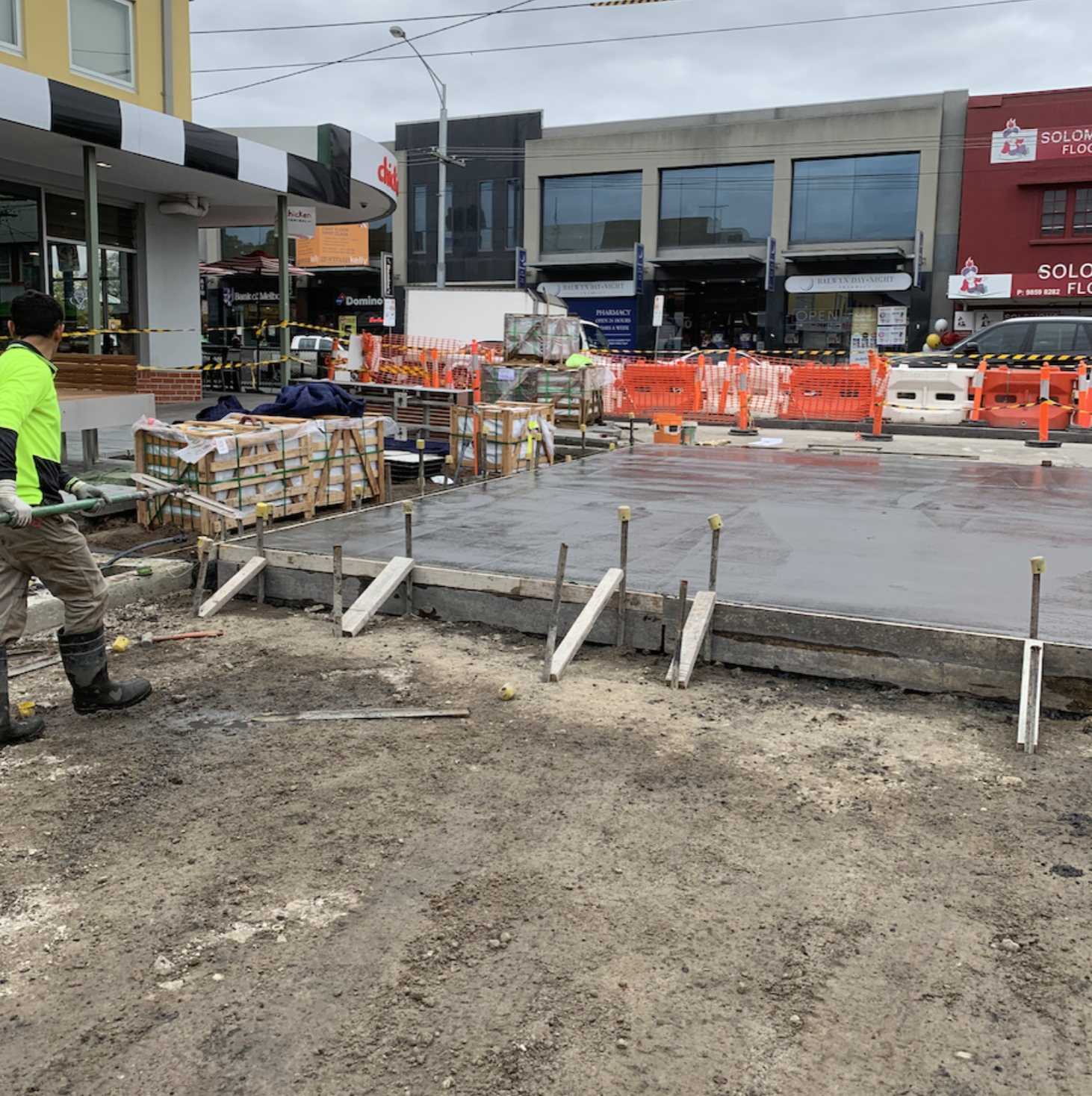 Construction workers are pouring concrete on a city street, surrounded by orange safety barriers and construction equipment, with storefronts and parked cars in the background.