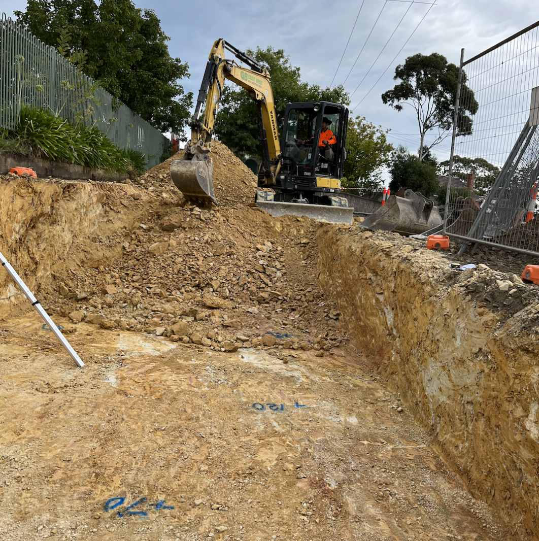 Construction site with a small excavator digging in the ground. The excavator operator is wearing an orange safety vest. There are trees and a fence in the background.