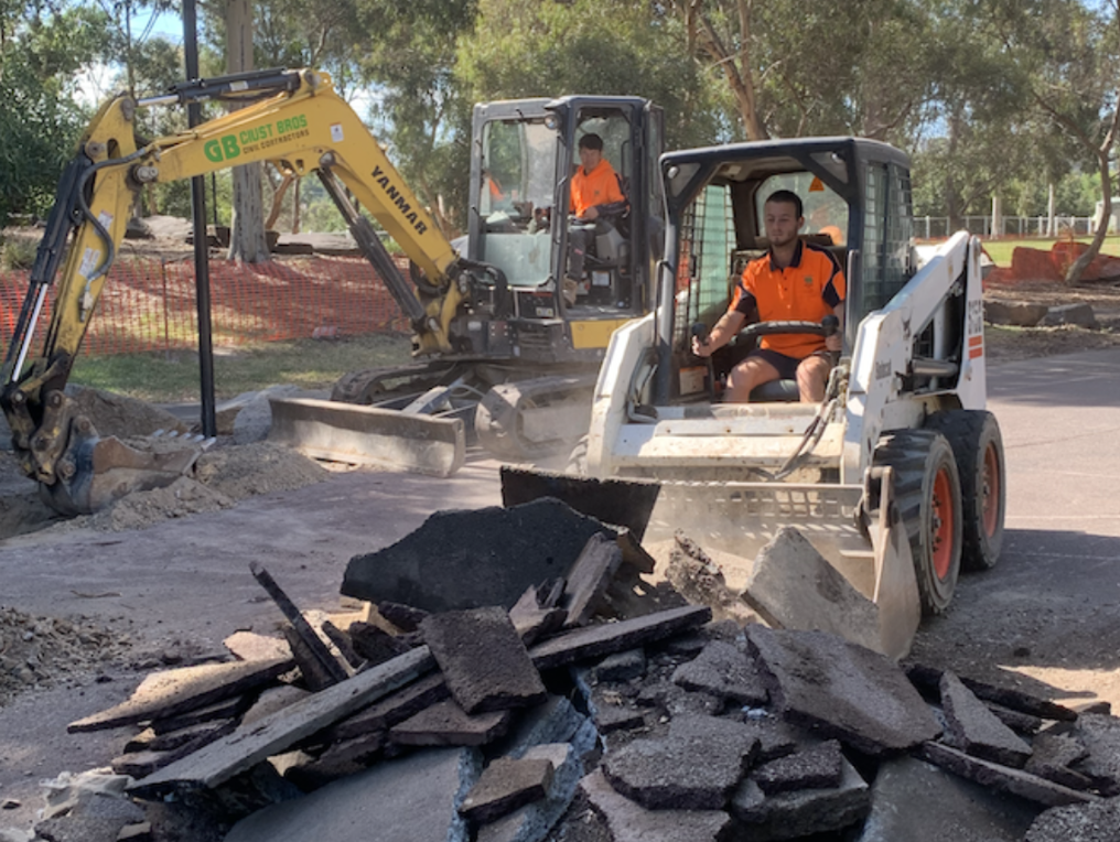 Two construction workers operating skid-steer loaders on a road under repair, with debris and broken asphalt in the foreground, trees and orange safety fencing in the background.