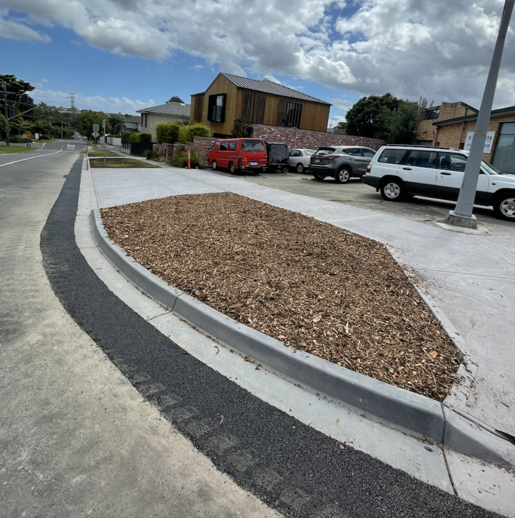 Sidewalk and curb with a landscaped area filled with wood chips, next to parked cars and residential buildings under a partly cloudy sky.