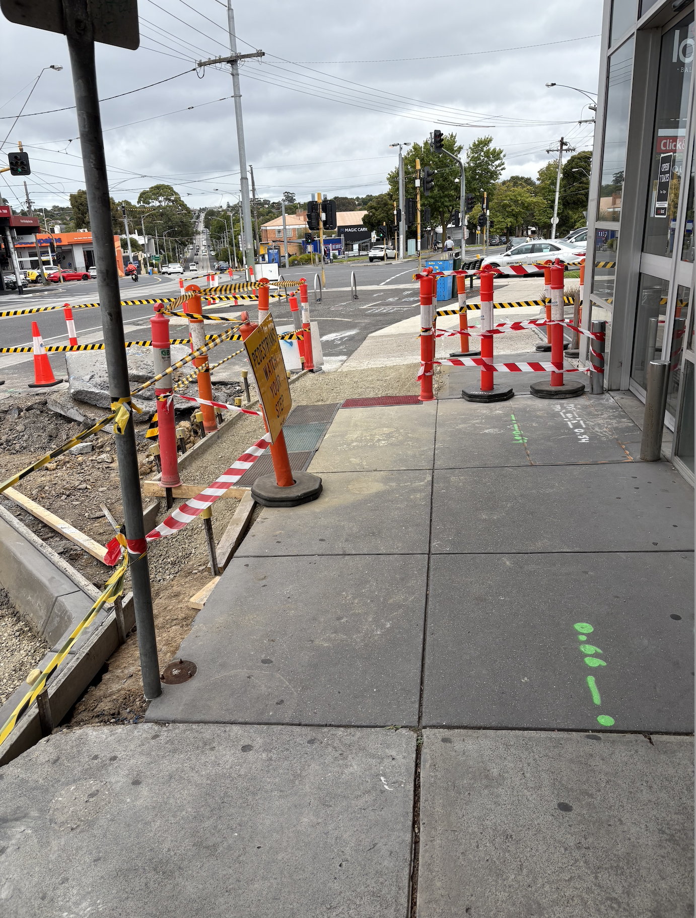 Sidewalk and street intersection with construction barriers, cones, and caution tape blocking part of the sidewalk and street, with traffic signals and cars in the background.