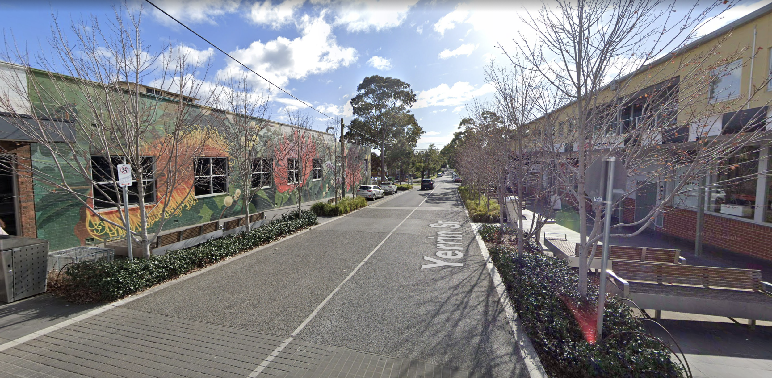 A street scene with trees lining the sidewalk, a colorful mural on the building on the left, benches on the right sidewalk, parked cars, and a blue sky with clouds.