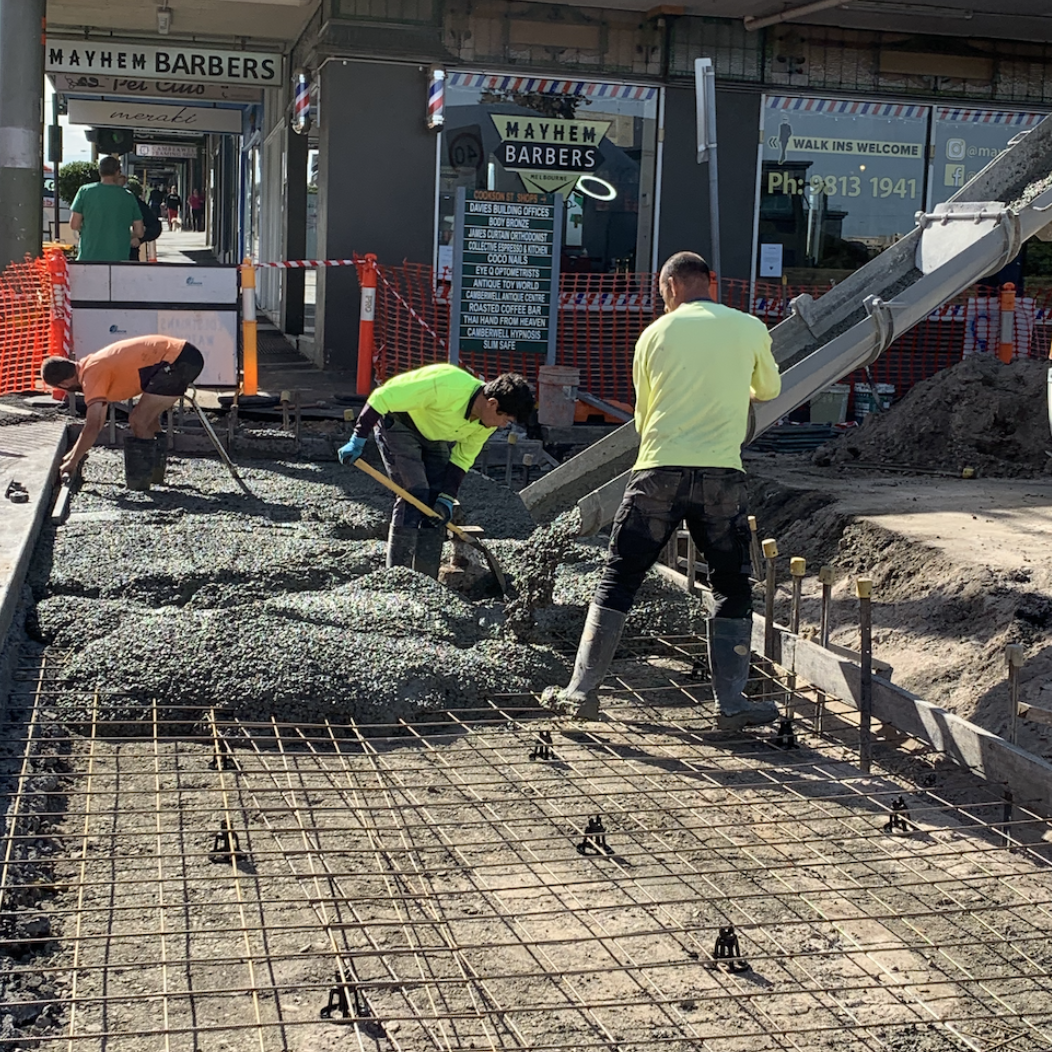 Construction workers pouring concrete on a sidewalk with rebar framework in front of a barber shop called Mayhem Barbers.