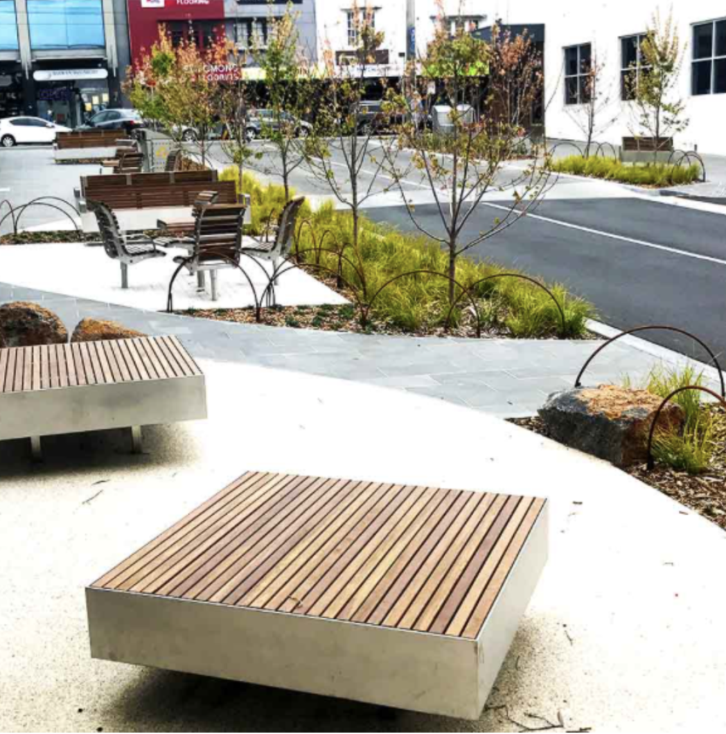 Urban park with modern benches and small trees along a sidewalk, with a street and buildings in the background.