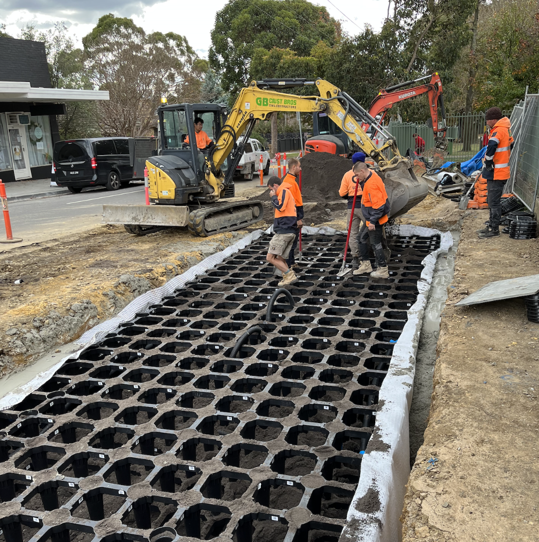 Construction workers in orange and black safety gear laying a grid of plastic foundation blocks on a dirt surface, with excavators and parked cars in the background.