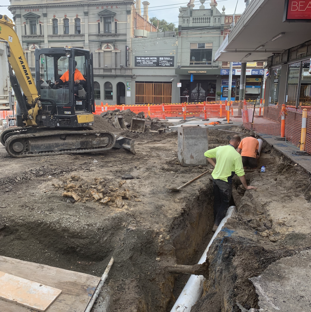 Construction workers install a pipe underground at a street excavation site with a small excavator and orange safety barriers in an urban area with buildings and businesses in the background.