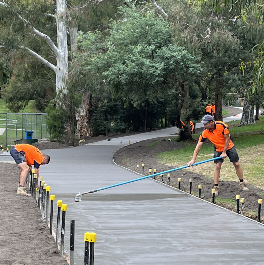 Workers in orange shirts finishing a concrete sidewalk in a park with trees and a winding path.