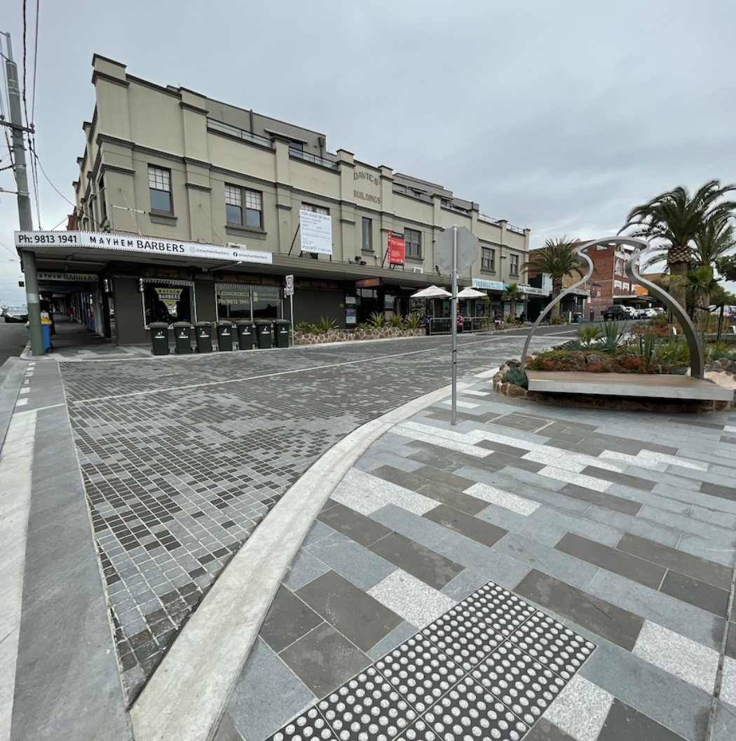A street scene with a building labeled 'Davies Buildings,' a barber shop called 'Mayhem Barbers,' palm trees, and a modern sculpture within a small garden area in an urban commercial zone.