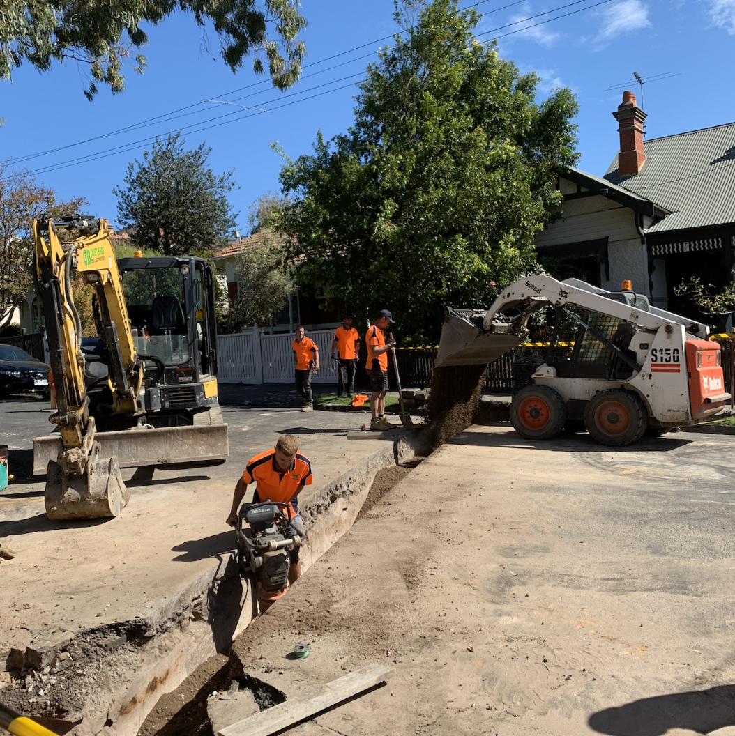 Construction workers operating heavy equipment, including an excavator and a skid-steer loader, on a street with a partially dug trench for roadwork or utility repairs, and a row of trees and houses in the background.