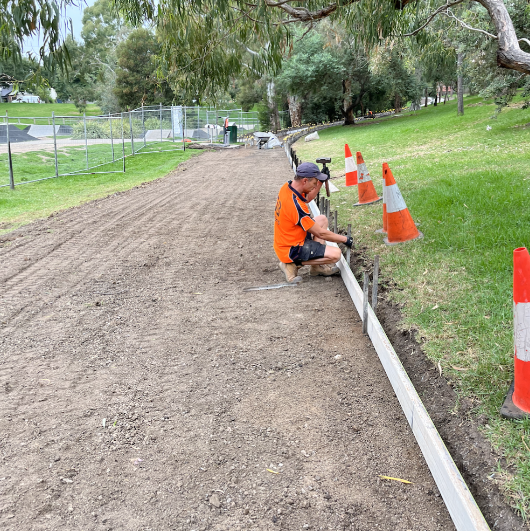 A worker in an orange shirt and cap installing a wooden board along a dirt pathway in a park, with orange and white traffic cones and trees in the background.