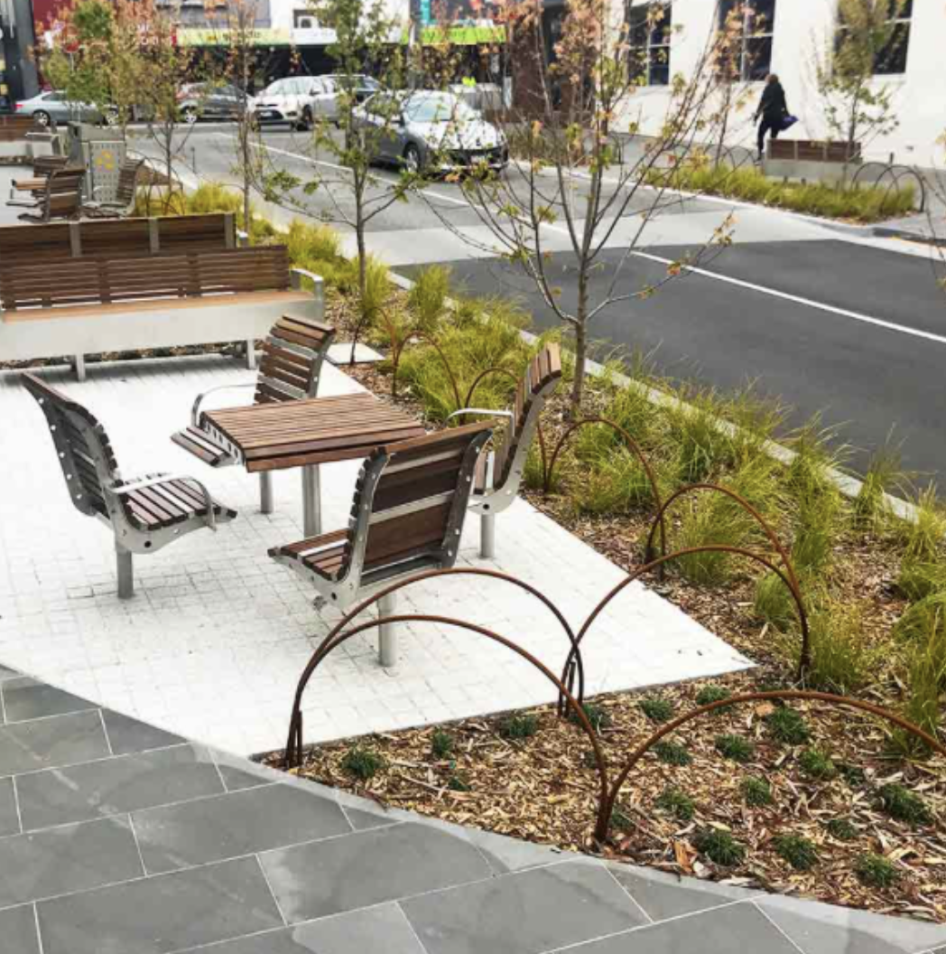 Outdoor seating area with wooden benches and tables, surrounded by small trees, plants, and decorative garden arches, with a parking lot and a person walking in the background.