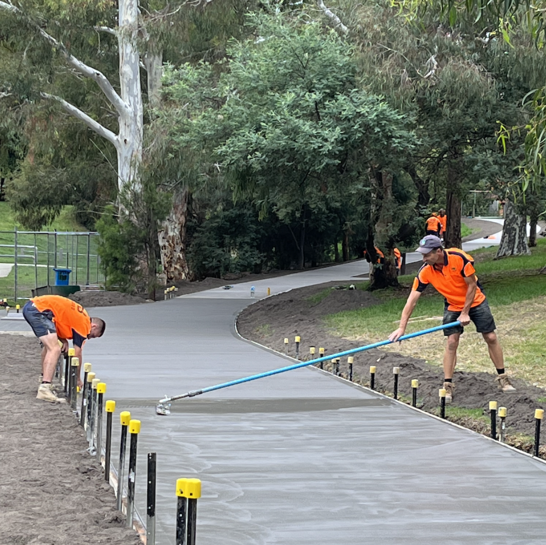 Construction workers in orange shirts pouring and smoothing concrete on a paved pathway in a park, surrounded by trees.