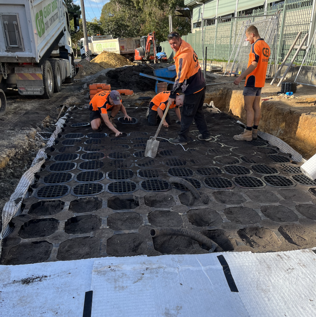 Construction workers installing a permeable grid on a sidewalk or driveway, with cars, trucks, and construction equipment in the background.