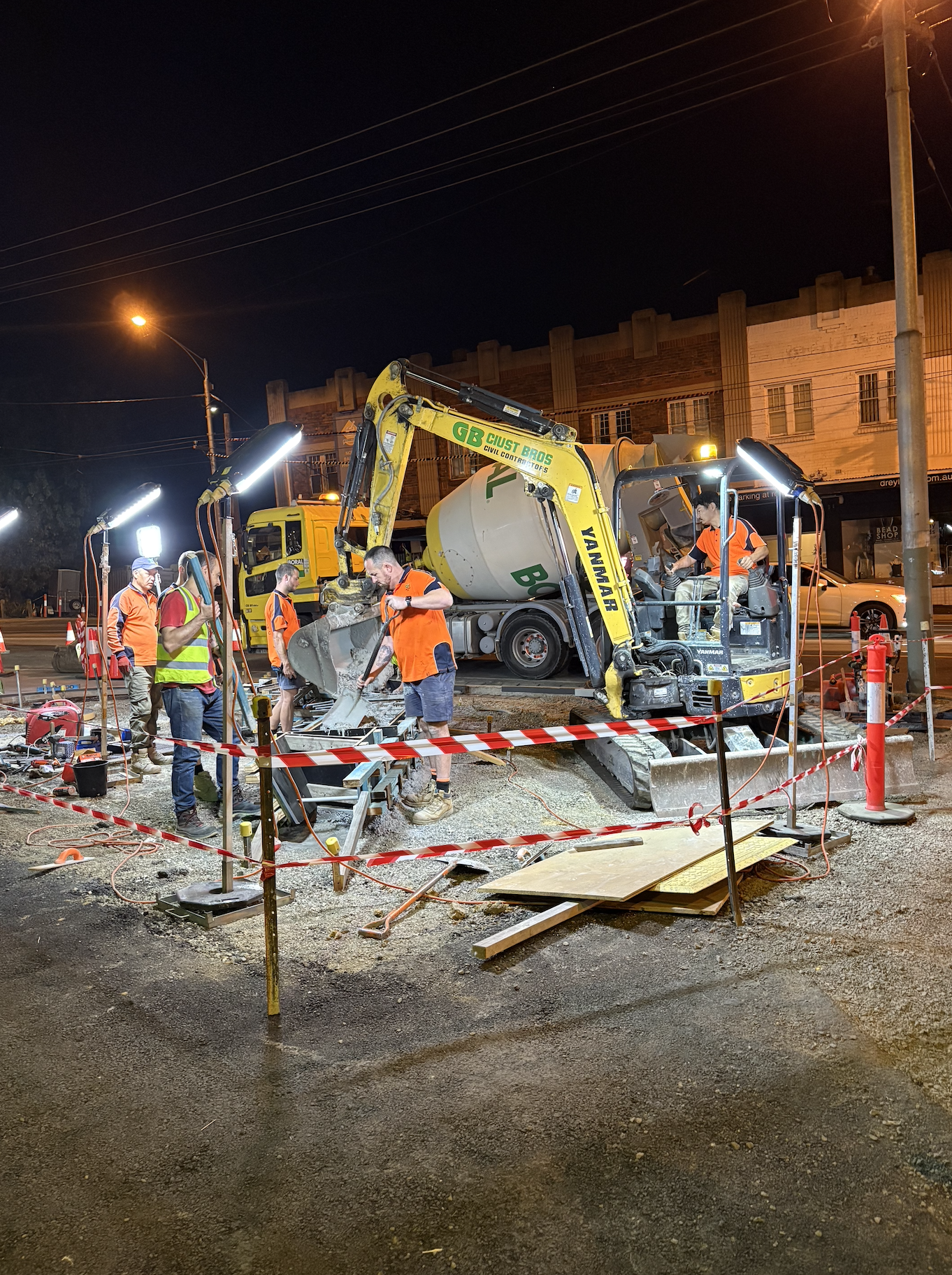 Workers at a nighttime construction site using a small excavator to break up pavement, with construction tools, equipment, and safety barriers around.