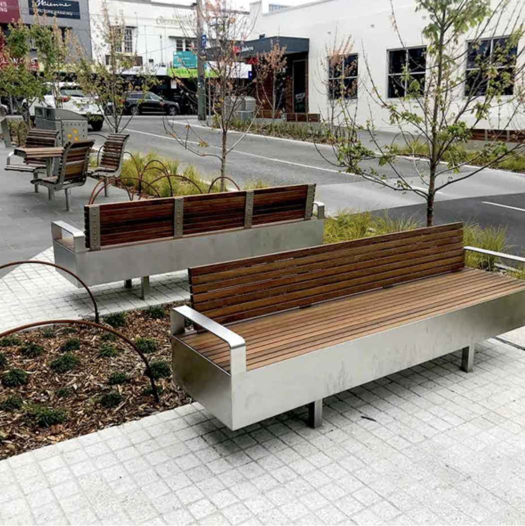 Empty outdoor benches along a city sidewalk with young trees and greenery, with a street and buildings in the background.