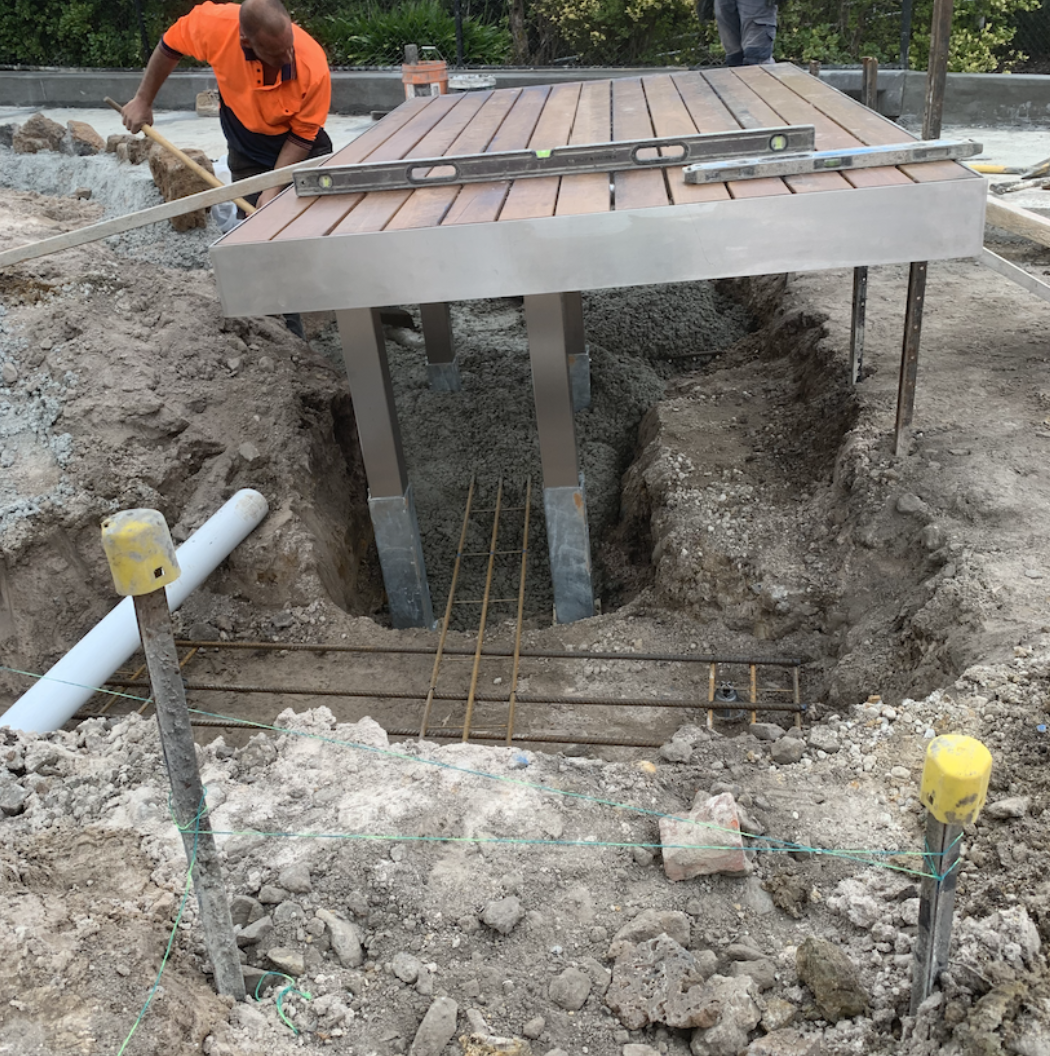 Construction workers are building a platform with a wooden deck supported by metal beams over a trench with concrete and rebar, with construction tools and pipes present on the site.