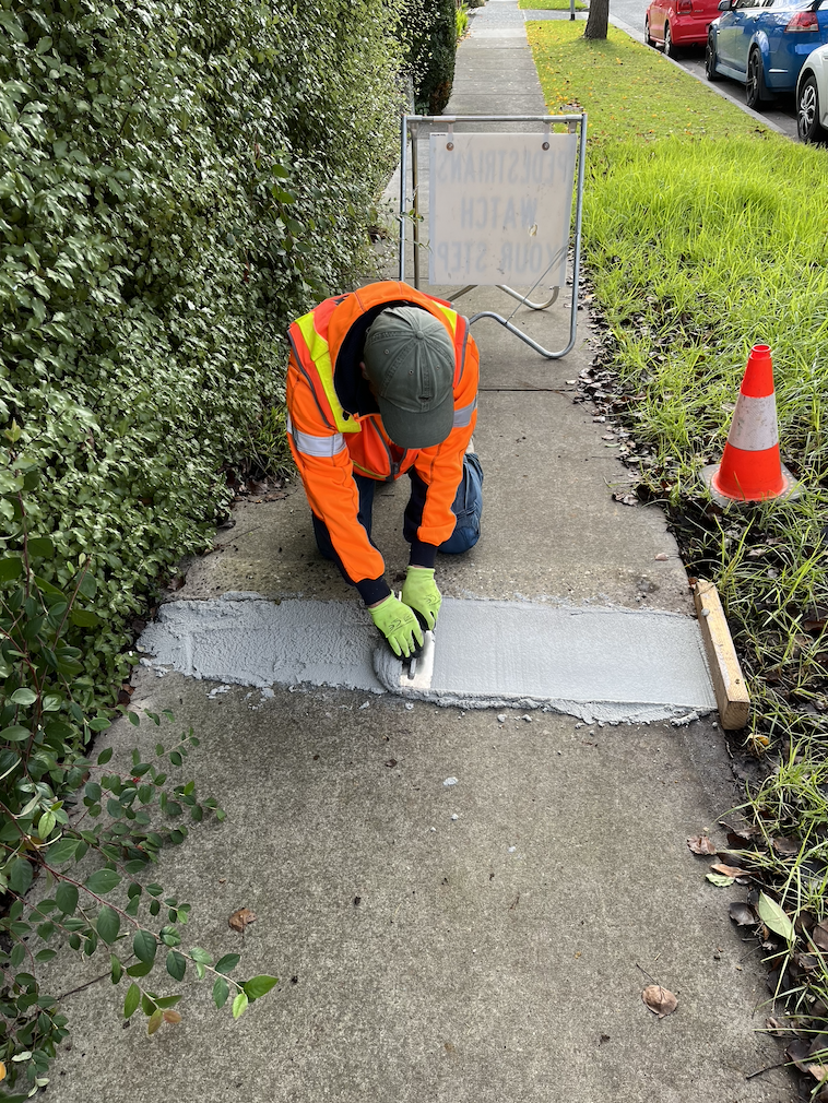Worker in orange safety jacket applying concrete patch on sidewalk with trowel, nearby traffic cone and construction barrier.