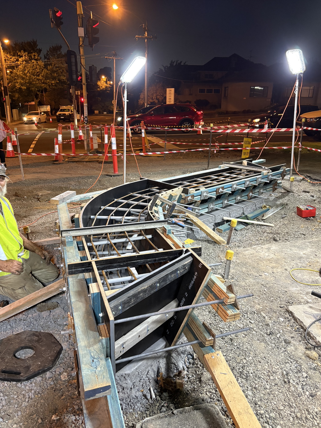 Nighttime construction site with steel framework, wooden forms, and construction worker in a yellow safety vest, set against a street with traffic, traffic lights, and houses in the background.