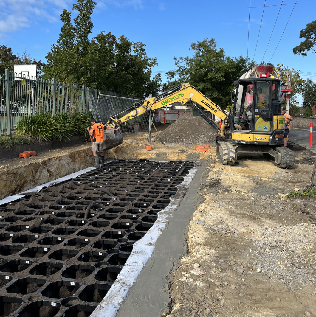 Construction site with workers installing a foundation, using a mini excavator and concrete grid, on a clear day with trees and a blue sky in the background.
