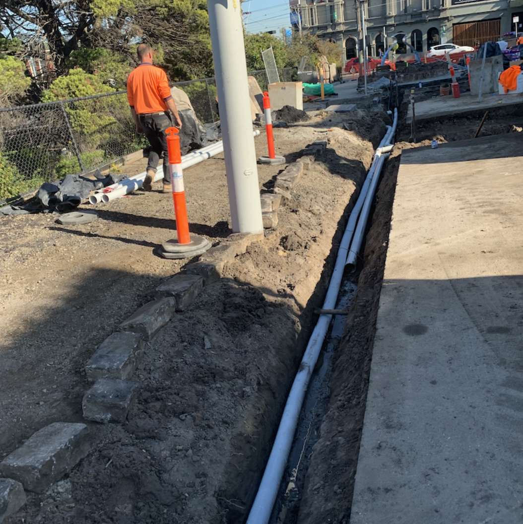 Construction workers installing underground pipes along sidewalk lined with construction cones and barriers, with buildings and parked cars in the background.