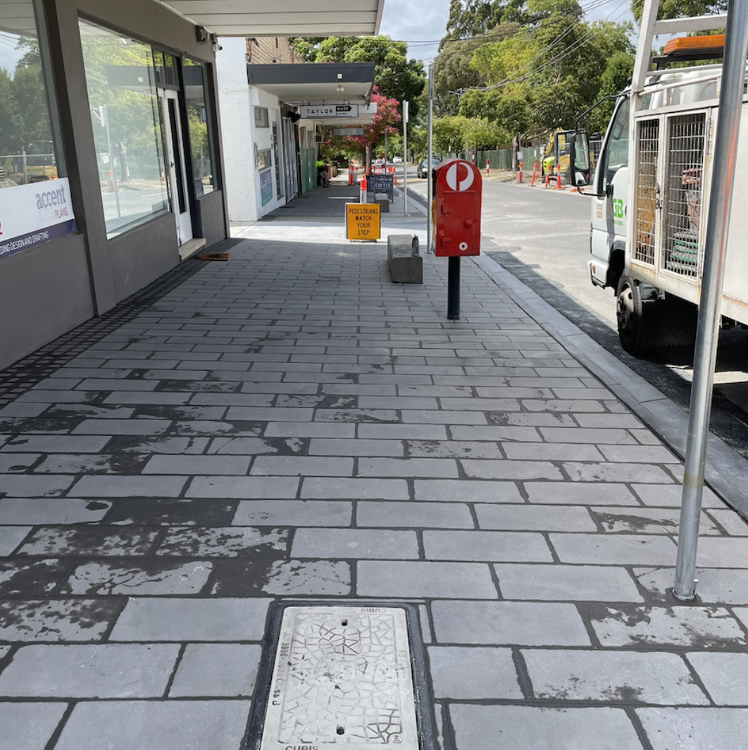 City sidewalk with shopfronts, a red mailbox, a sign that says 'Pedestrians Watch Your Step', and a construction truck parked on the street, with trees in the background.