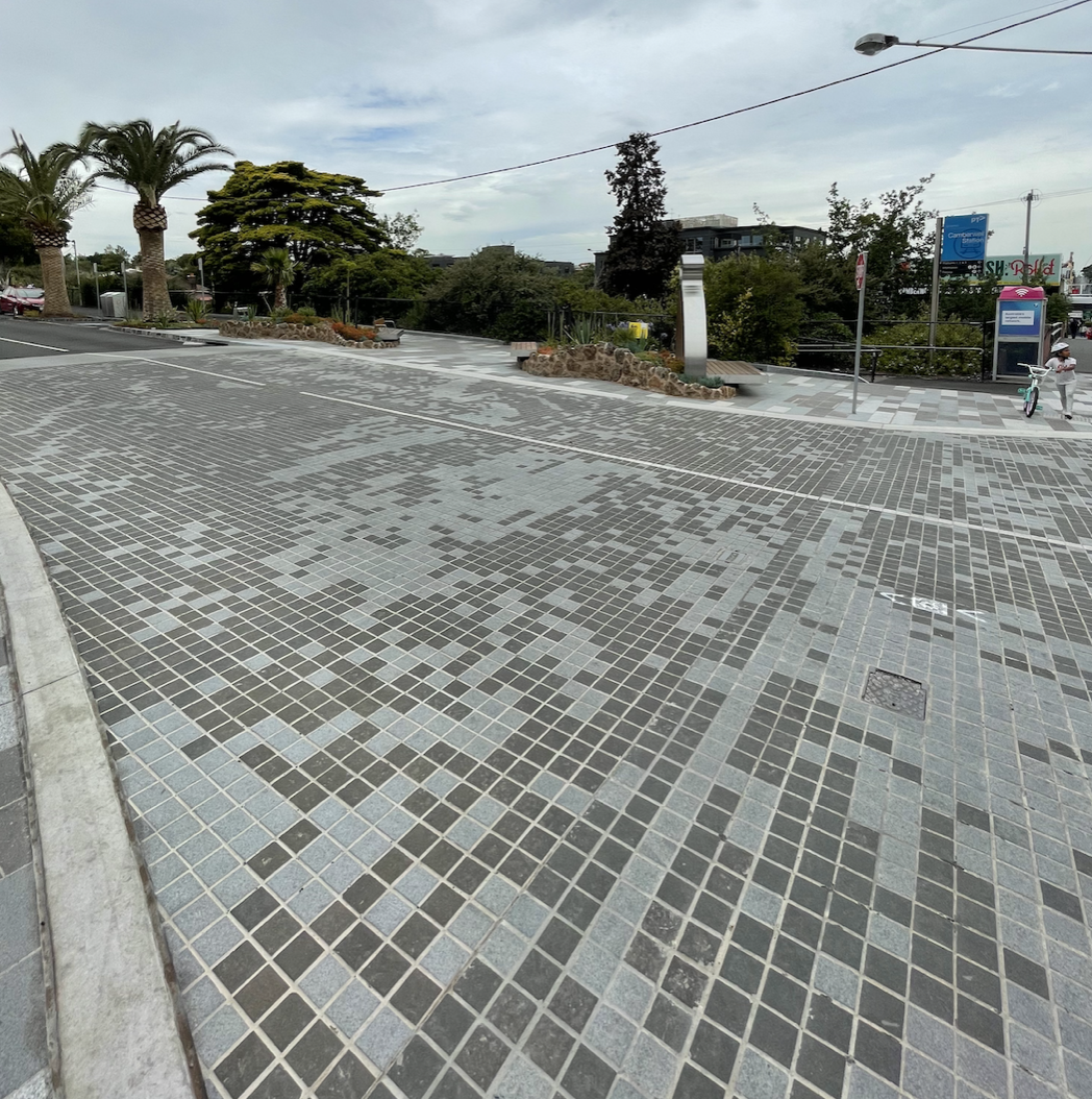 A paved urban plaza with small square tiles in shades of gray and black, surrounded by trees, poles, and a few people in the background.
