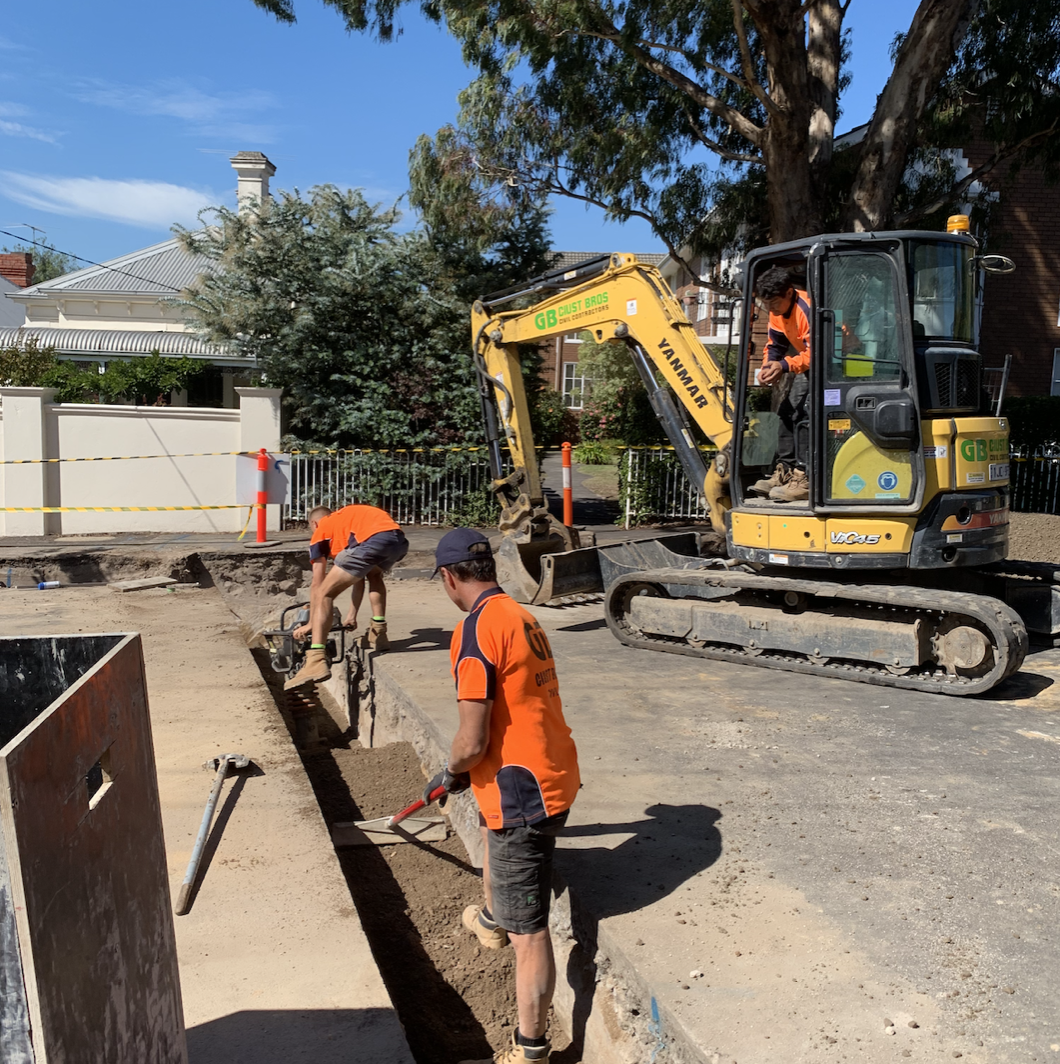 Construction workers in orange shirts working on a sidewalk with a small excavator.