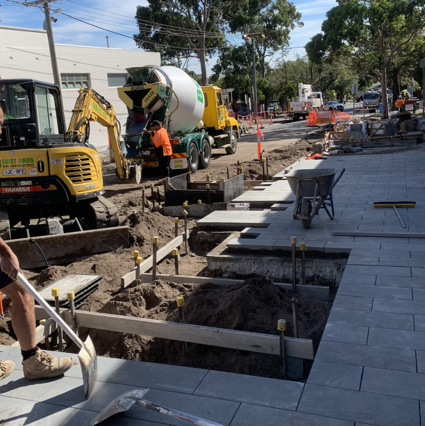 Construction workers laying paving stones on a sidewalk, with construction equipment and vehicles on the street, and workers and vehicles in the background.