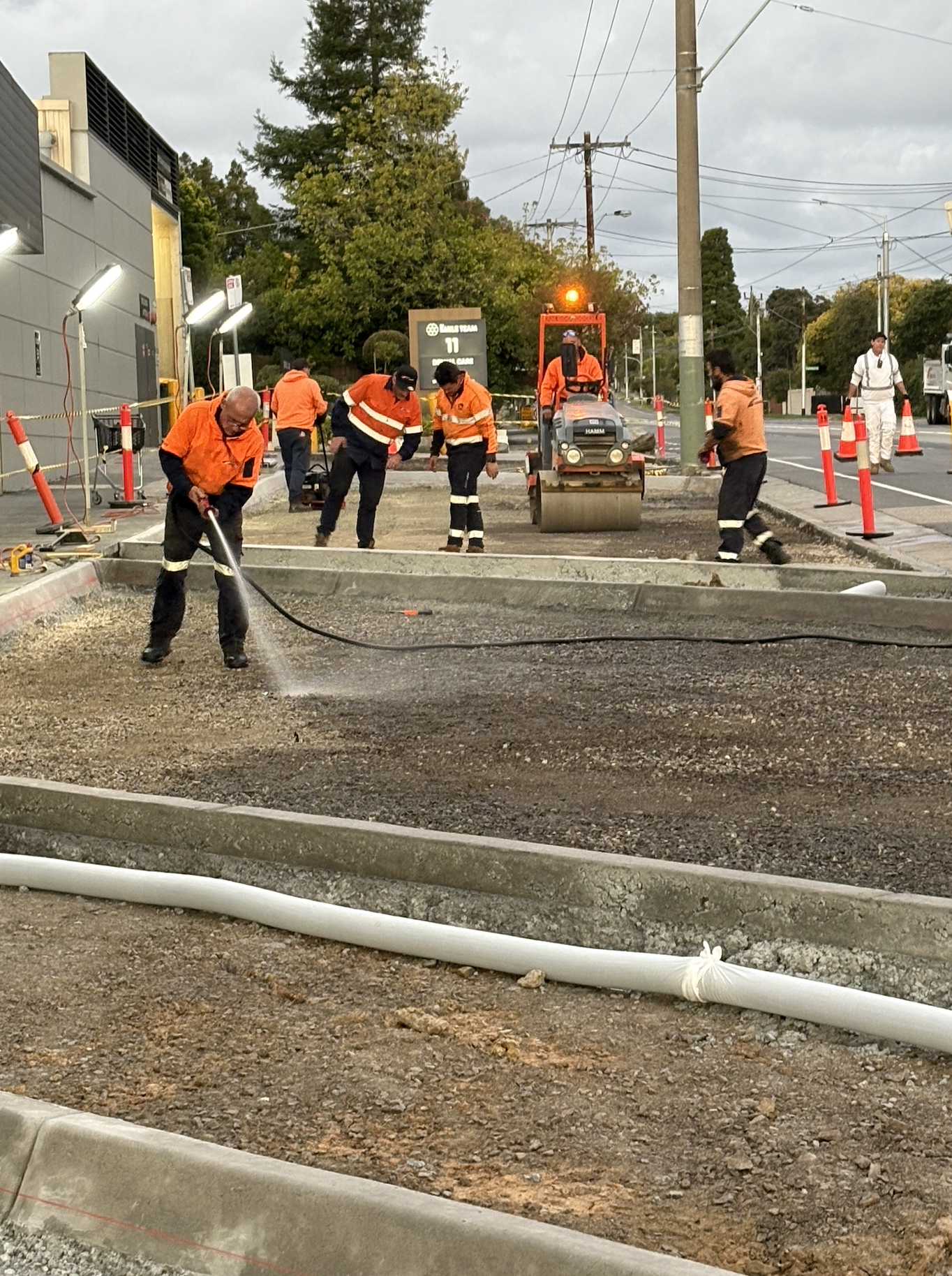 Construction workers paving a road, with some using power tools and others operating machinery, while traffic cones and safety equipment surround the work site.