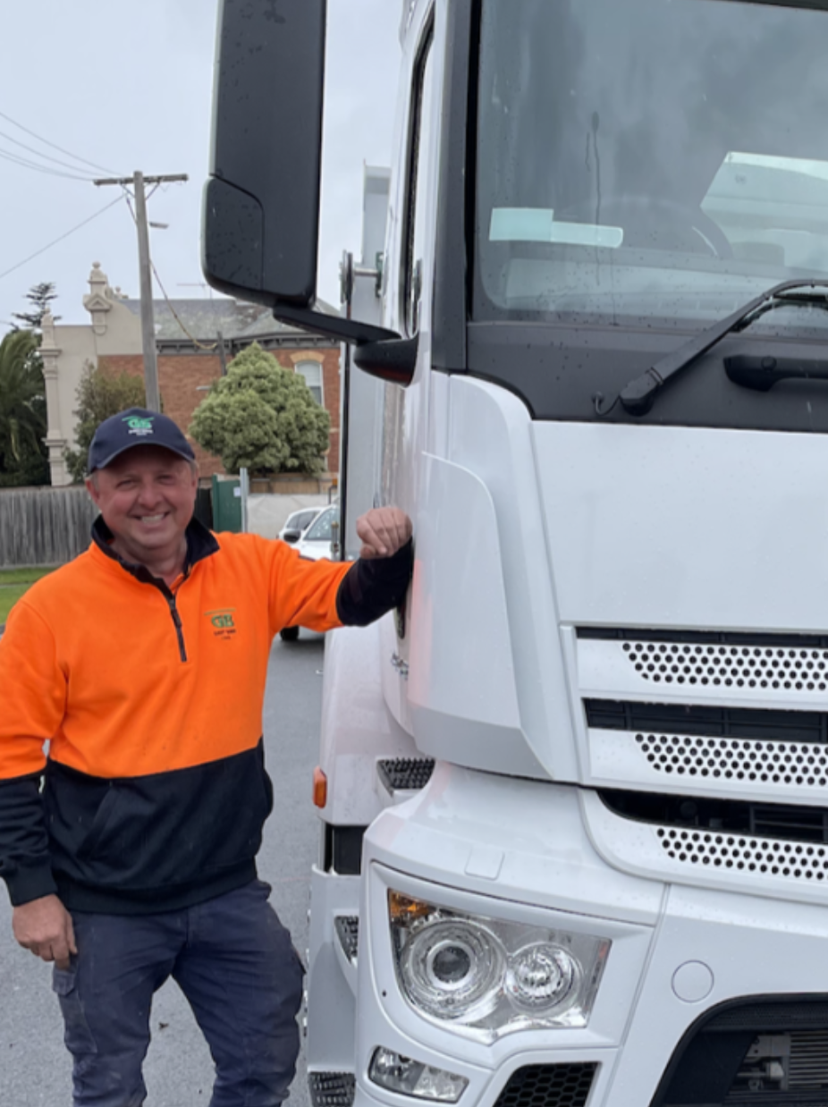 A smiling man wearing an orange and black jacket and a blue cap standing next to a large white truck on a street with houses in the background.
