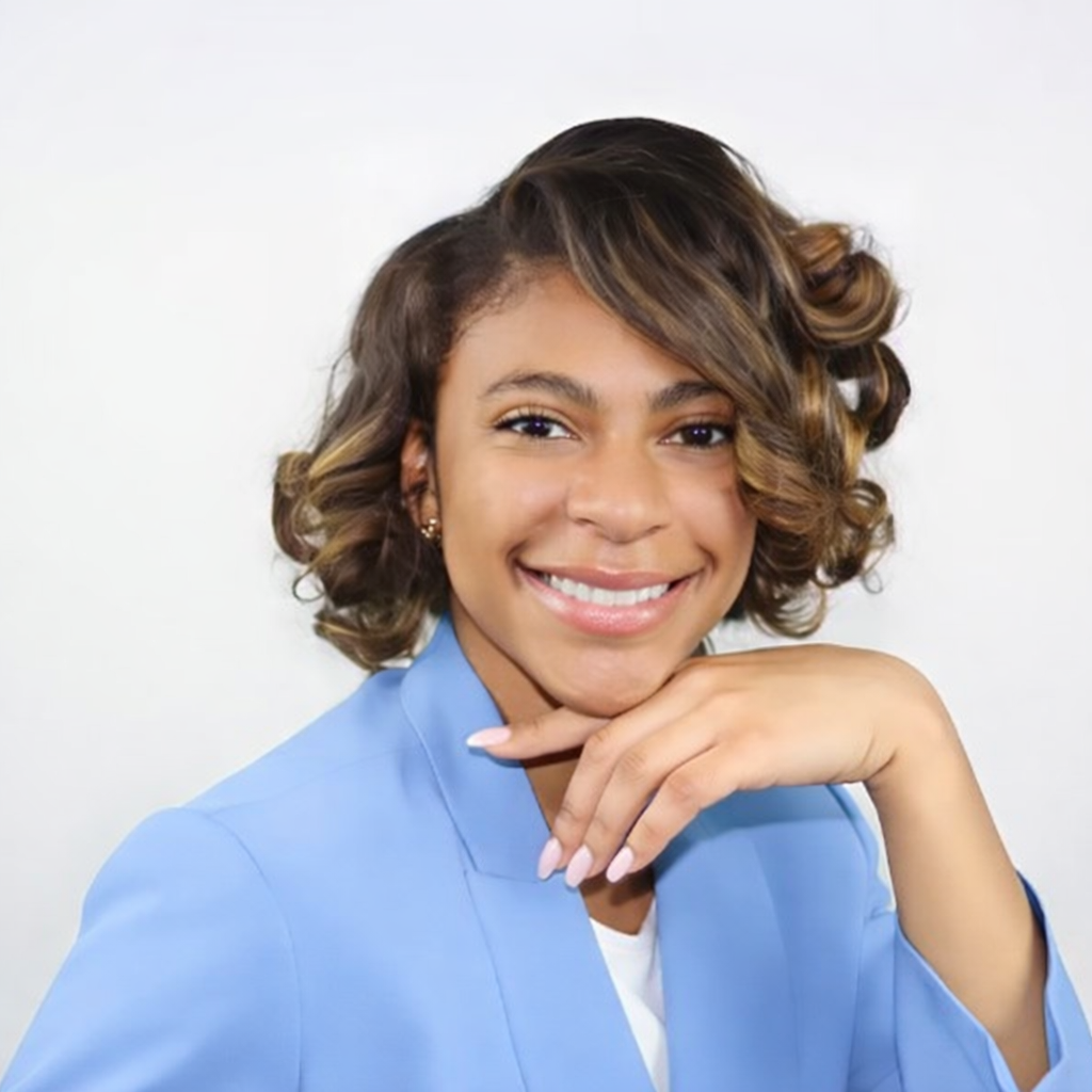 A young woman with curly hair styled with highlights, smiling and resting her chin on her hand, wearing a light blue blazer and white top against a plain white background.