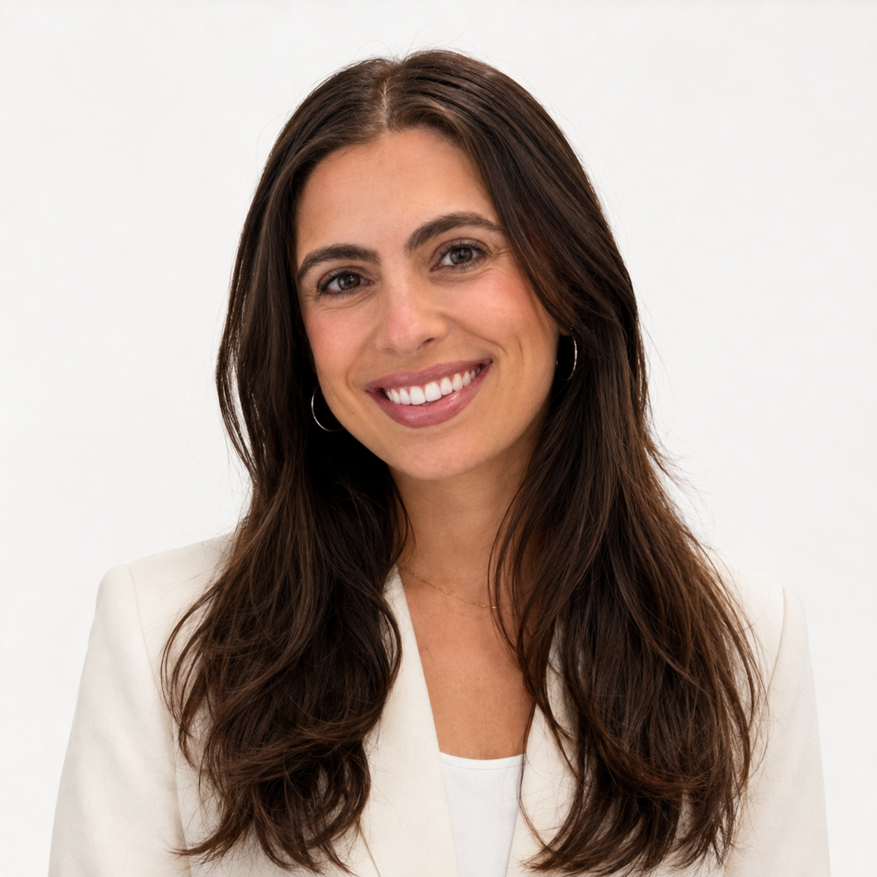 A woman with long brown hair smiling, wearing a white blazer, hoop earrings, and a delicate necklace against a plain white background.