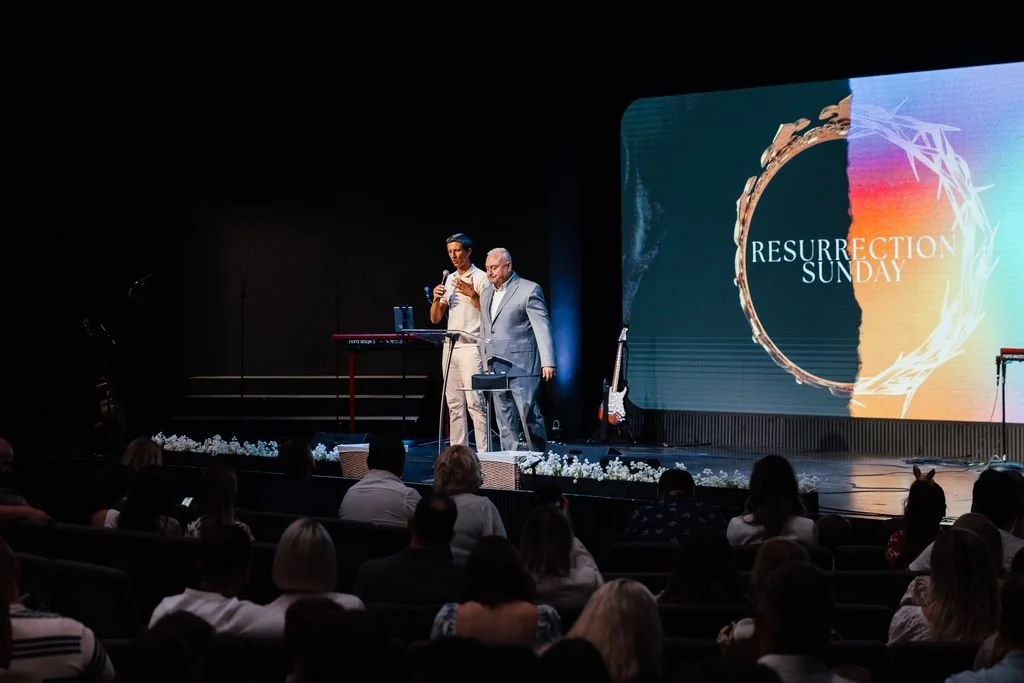 Two men on stage at a church or event, one speaking into a microphone, the other looking at a phone, with a digital screen in the background displaying 'Resurrection Sunday.' An audience is seated in front of them.
