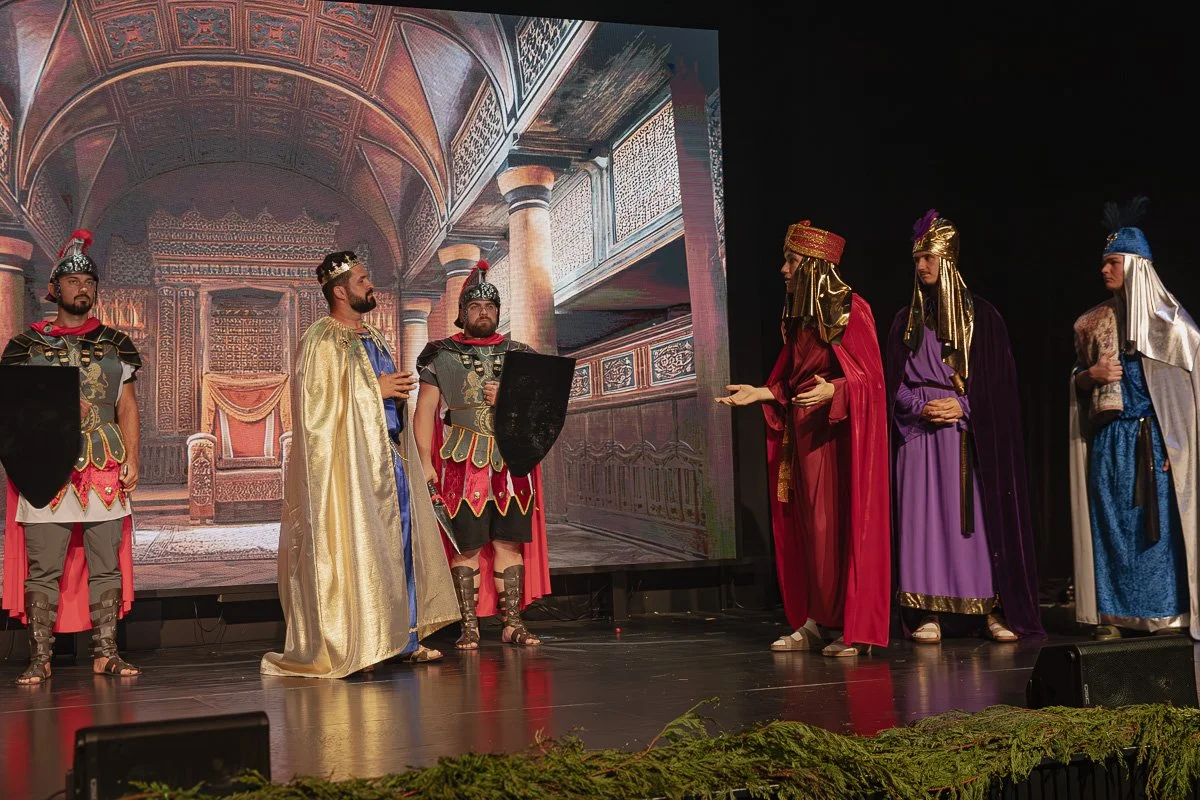 A theatrical performance on stage with actors dressed as Roman or biblical characters, standing in front of a large backdrop depicting an ornate palace interior.