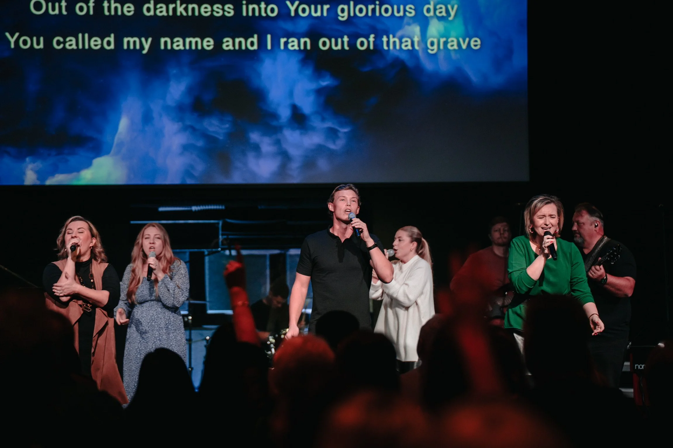 Group of six people singing and playing music on stage during a concert, with lyrics displayed on a large screen behind them.