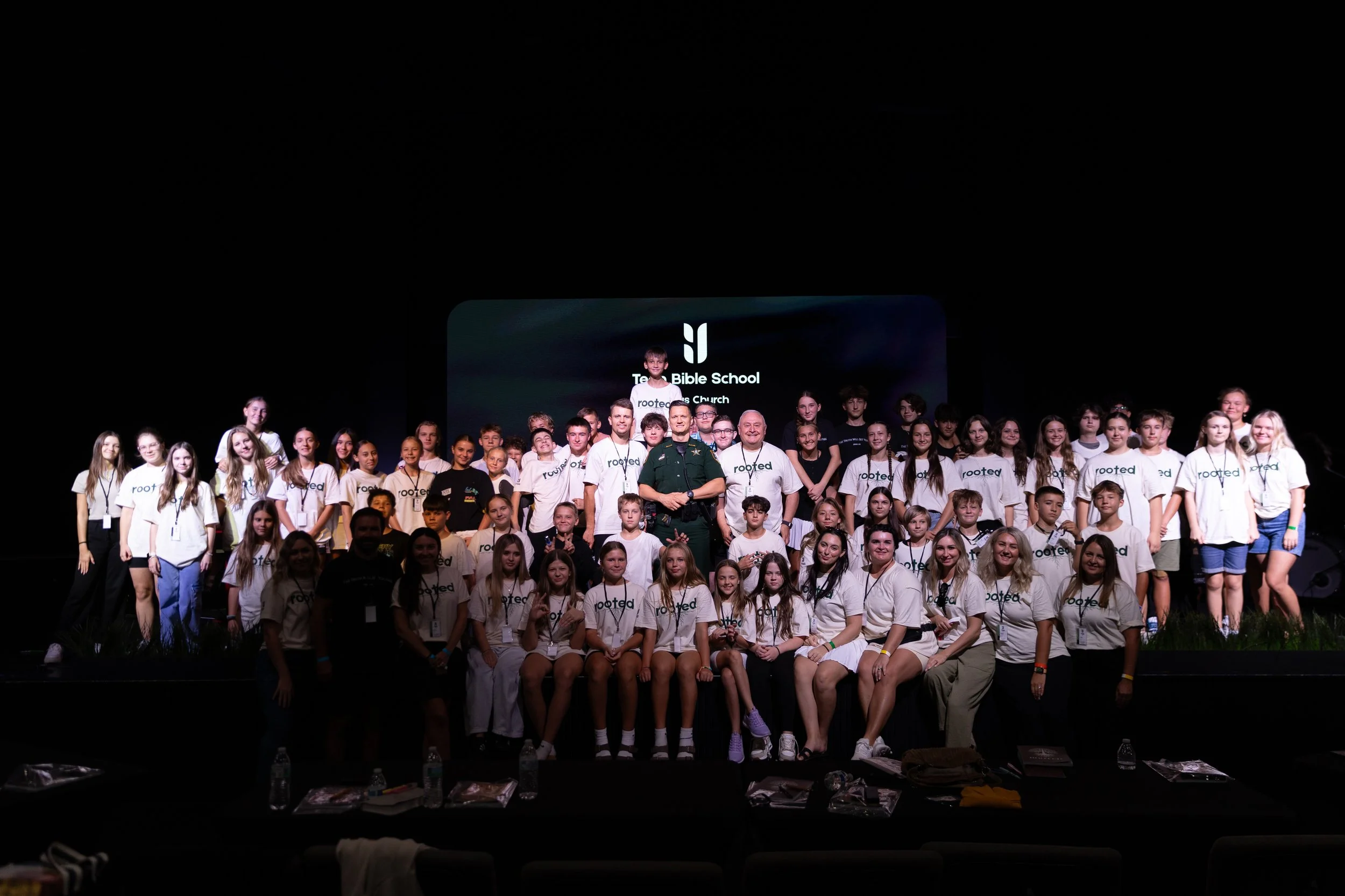 A large group of children and adults on stage at a church event, with a sign in the background reading 'Tree Bible School' and 'Rooted.' The group is diverse, with some wearing white 'Rooted' t-shirts and others in casual clothing, posing for a group photo in a dark theater setting.